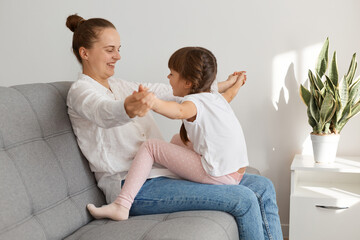 Loving mom laughing caressing with cute kid daughter sitting on gray sofa at home, happy mother and little child girl having fun, playing, enjoy funny moments and good time together.