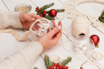 A woman decorates a transparent Christmas ball with macrame-style weaving,handmade decor in eco-style,hands close-up.Christmas, New Year and eco-friendly concept.Selective focus.