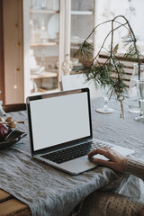 Wall Mural - Woman work on laptop computer with blank screen on linen table cloth with fir spruce bouquet, Christmas decorations. Elegant home office working space interior design. Freelancer works at home