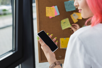 Wall Mural - cropped view of businesswoman with pink hair holding smartphone with blank screen near board with sticky notes.