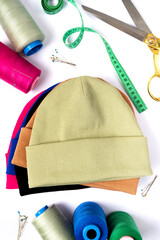 Wall Mural - Top view of a stack of insulated stylish beanie hats in different colors with scissors, threads and needles. Tailoring concept. Vertical photo