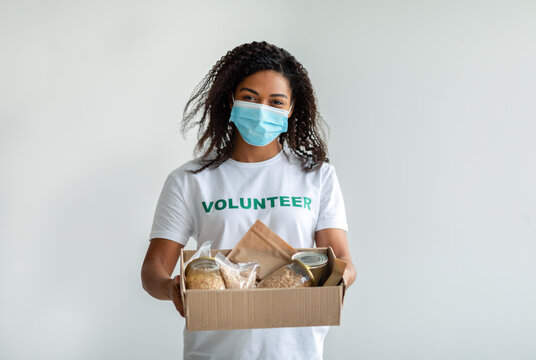Young african american female volunteer in medical face mask holding donations box, standing on white studio background