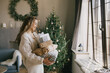 © polinaloves - Young beautiful woman holding gift boxes and presents in a cozy room with Christmas tree on background. Holiday preparations.