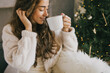 © polinaloves - Young woman drinking hot coffee or cocoa sitting on couch in cozy room with Christmas tree. Winter holidays.