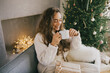 © polinaloves - Young woman drinking hot coffee or cocoa sitting on couch in cozy room with Christmas tree. Winter holidays.