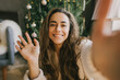 © polinaloves - Young woman waving her hand, video chatting with family and friends on Christmas.