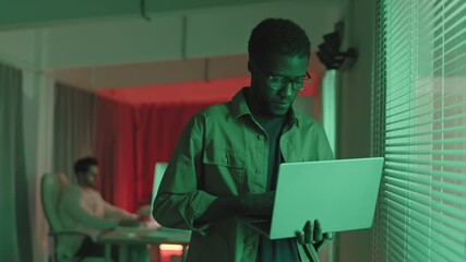 Wall Mural - waist-up slowmo shot of young african-american programmer typing on laptop standing in dark office w