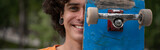 joyful young man obscuring face with skateboard while looking at camera, banner.
