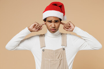 Wall Mural - Young african man in Santa Claus red Christmas hat cover ears with hands fingers do not want to listen scream isolated on plain pastel beige background studio. Happy New Year 2022 celebration concept.