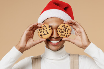 Wall Mural - Young cheerful fun african man 20s in Santa Claus red Christmas hat cover eyes with cookies biscuits isolated on plain pastel beige background studio portrait. Happy New Year 2022 celebration concept.