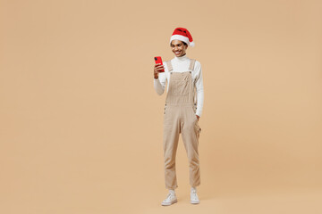 Wall Mural - Full body young african man 20s in Santa Claus red Christmas hat hold in hand use mobile cell phone isolated on plain pastel beige background studio portrait. Happy New Year 2022 celebration concept