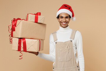 Young courier african man 20s wear Santa Claus red Christmas hat hold many present boxes with gift ribbon bow isolated on plain pastel beige background studio. Happy New Year 2022 celebration concept.