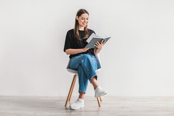 Stay home hobbies. Positive young woman reading favorite book, sitting on chair against white studio wall, full length