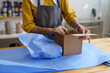 © DimaBerlin - Closeup of female ceramist packing crockery in box for delivery. Woman in apron wrap pottery in cardboard for shipping. Handcraft artist prepare handmade present. E-commerce and small business concept