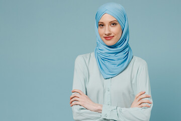 Wall Mural - Young smiling confident arabian asian muslim woman in abaya hijab hold hands crossed folded isolated on plain blue color background studio portrait. People uae middle eastern islam religious concept.