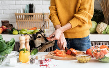 Front View Of Female Hands Making Salad And Giving A Piece Of A Vegetable To A Dog