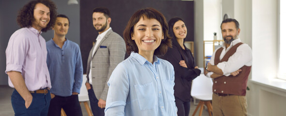 Wall Mural - Smiling young woman standing in front of group of coworkers. Banner with portrait of happy female business leader together with team of employees and colleagues in blurred office background behind