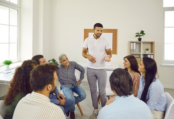 Wall Mural - Handsome young business coach talking to group of people during meeting in modern office. Team of happy young and senior employees listening to friendly manager or team leader