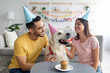 Millennial multiracial couple in party hats celebrating pet dog's birthday with small festive cake at home