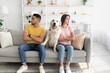 Unhappy offended young diverse couple looking in opposite directions, sitting on sofa with cute dog between them