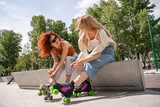 cheerful woman in sunglasses tying laces on roller skate on border near friend.