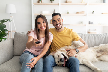 Poster - Happy young interracial couple sitting on couch with their funny dog and watching TV at home