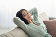 © Prostock-studio - Lazy weekend at home. Charming Indian woman resting with hands behind head on cozy couch at living room