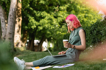 Wall Mural - side view of happy woman with pink hair holding paper cup and using laptop on grass.