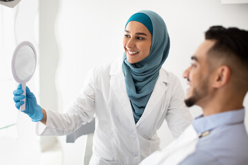 Wall Mural - Muslim Dentist Woman Holding Mirror, Showing Teeth Treatment Result To Male Patient