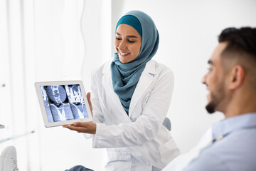 Wall Mural - Muslim Female Dentist Holding Digital Tablet, Explaining Treatment Process To Male Patient