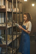 © pressmaster - Young serious craftswoman in apron scrolling in digital tablet while standing by shelves with handmade earthenware in storage room