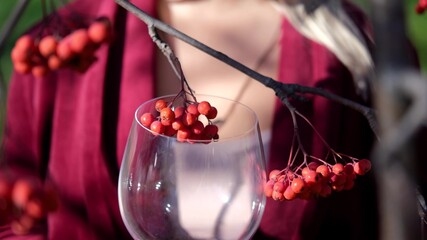 Smiling beautiful blond woman stands with a glass of wine in the sun on the background of rowan branches. Autumn atmosphere and mood