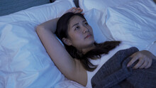 Sleepless Asian Girl Covered With Blanket Resting In Her Dark Bedroom Putting One Hand Back Of Head And Staring Upward In Contemplation.