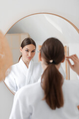 Wall Mural - Young woman applying natural essential oil with pipette on her face near mirror