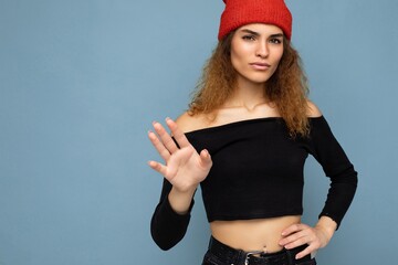 Wall Mural - Photo of pretty beautiful attractive cool young curly dark blonde woman standing isolated over blue background wall with copy space for text wearing everyday trendy and stylish black top and red hat