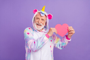 Sticker - Portrait of attractive bearded cheerful grey-haired man in kigurumi holding heart amour isolated over violet purple color background