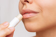 © Марина Демешко - Cropped shot of a young caucasian beautiful woman applying a hygienic lipstick on her lips on a gray background. Moisturizing chapstick for dry lips