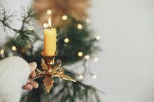 Atmospheric Hygge Winter Home. Hand In Cozy Sweater Holding Vintage Candlestick With Burning Candle On Background Of Warm Lights, Fir Branches, Sweden Star In Festive Scandinavian Room