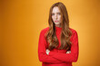 Suspicious intense and defensive ginger girl standing in passive-aggressive pose pouting and frowning looking with disbelief and disdain at camera, offended against orange background