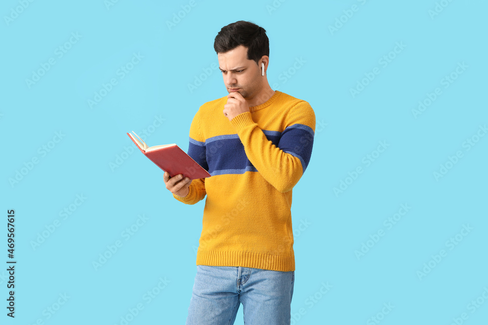 Frowning man with book and earphones on color background