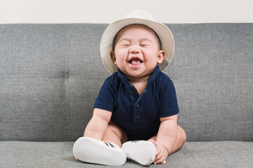 Adorable baby boy  first sitting on sofa. Cute infant Asian about 5-6 months old wearing blue t-shirt,white hat and shoes smiling and looking at camera.