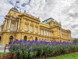 Croatian National Theater in Zagreb shown behind rows of beautiful purple flowers and greenery with blue sky and clouds in the background. 