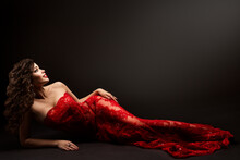 Sexy Brunette Woman In Red Lace Dress Lying Down Over Black. Fashion Model In Long Evening Gown With Curly Hair Looking Up Happy Smiling