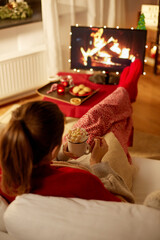Wall Mural - christmas, winter holidays and leisure concept - young woman watching tv with fireplace on screen and holding mug of marshmallow and whipped cream resting feet on table at cozy home