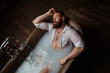 An athletic bearded man relaxes in a luxurious bath in a dark interior.