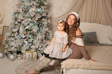 Wall Mural - beautiful young long-haired girl in a hat of sanat klaus in a knitted sweater in long leggings sits on the bed holding a little daughter near the christmas tree