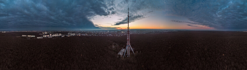 Aerial panorama after sunset, evening view on dark telecommunication tower antenna silhouette in center of forest and scenic cloudy sky. Kharkiv, Ukraine