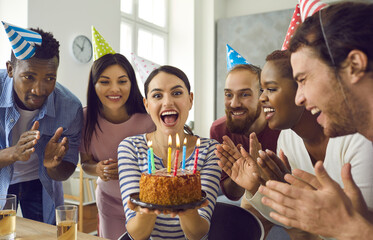 Close up of a birthday girl surrounded by her friends makes a wish and is going to blow out the candles on the cake. Group of cheerful young multiracial people in conical party hats applaud.