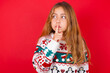 © Jihan - little kid girl wearing knitted sweater christmas over red background makes silence gesture, keeps index finger to lips makes hush sign. Asks not to share secret.