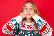 © Jihan - Happy little kid girl wearing knitted sweater christmas over red background keeps fists on cheeks smiles broadly and has positive expression being in good mood